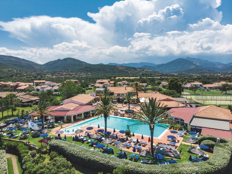 View of a resort with two pools, sun loungers, and mountains in the background under a cloudy sky.