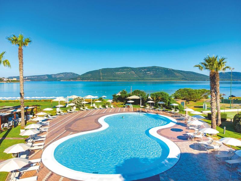 Hotelzwembad met ligstoelen, palmbomen en uitzicht op zee en bergen bij heldere lucht.