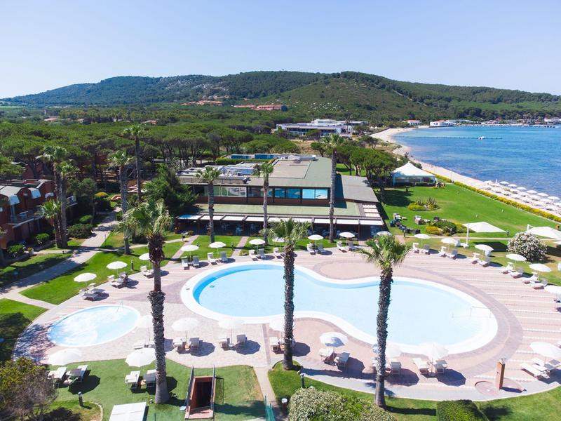 Hotelzwembad met parasols, palmbomen en uitzicht op zee en strand in heuvelachtig landschap.