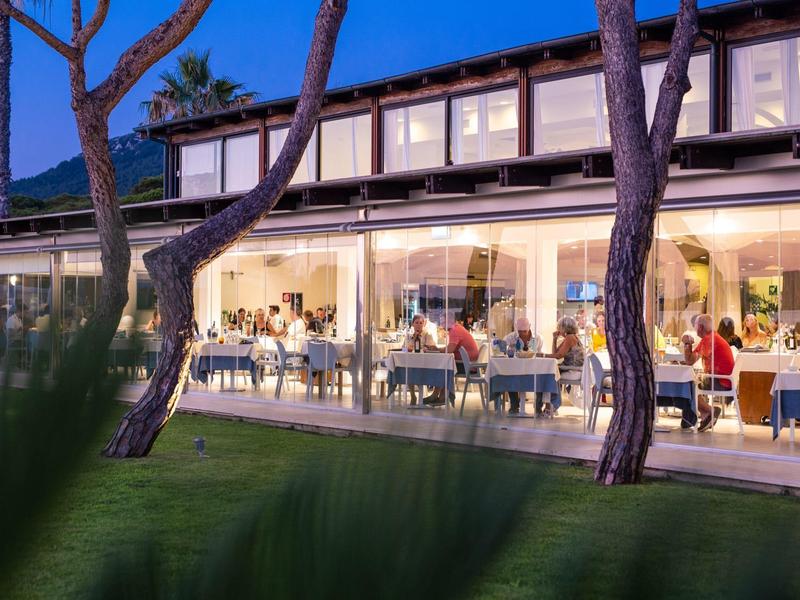 Modern hotel met verlicht restaurant en mensen die buiten dineren bij schemering.