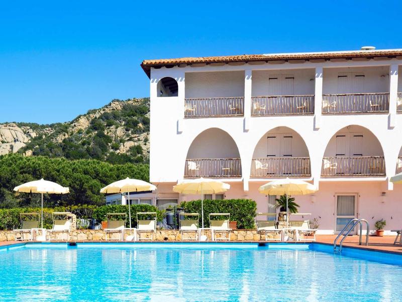 Light blue pool in front of a hotel building with arches and sun loungers under a clear blue sky.