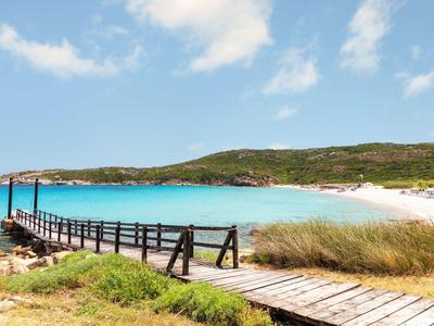 Holzsteg führt zu einem Strand mit türkisfarbenem Wasser und grünen Hügeln im Hintergrund.
