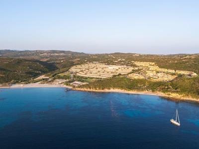 Küste mit blauem Meer und einem Segelboot, darüber Hügel mit Gebäuden unter klarem Himmel.