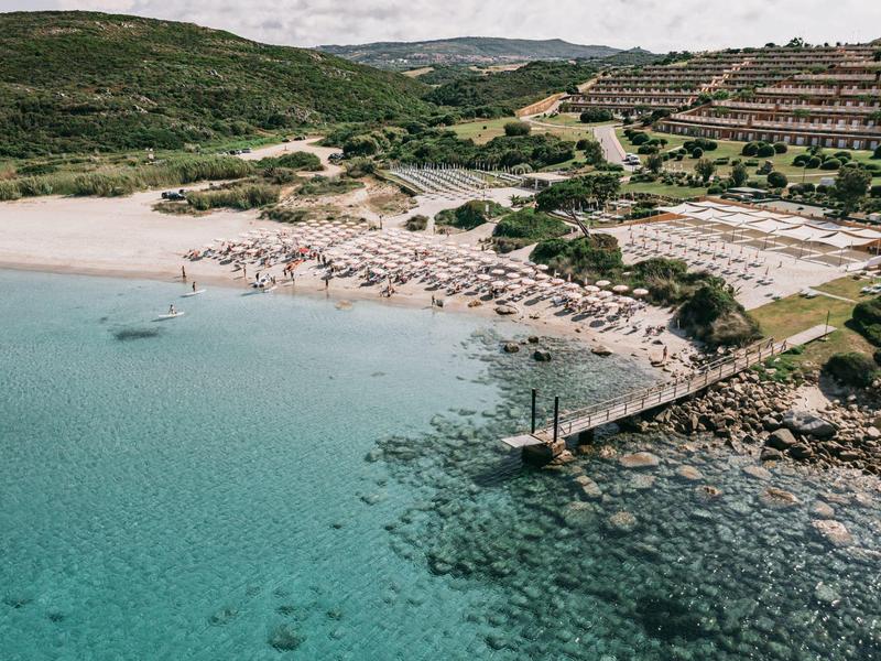 Türkisblaues Meer mit Sandstrand, Liegestühlen, grünen Hügeln und einem Steg ins Wasser.