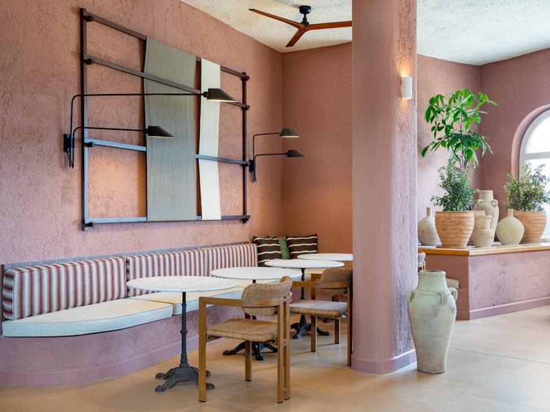 Gemütlicher Hotelmorgenbereich mit Sitzbank, Tischen, Stühlen und großen Fensterpflanzen.