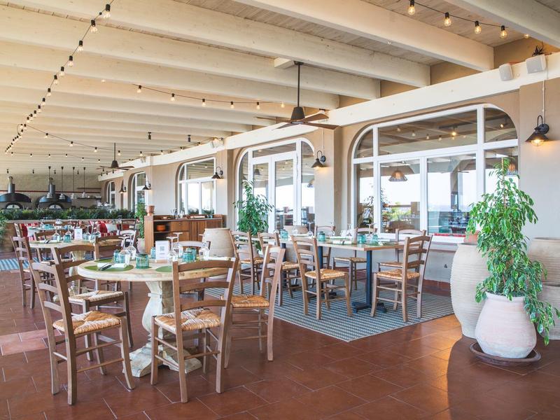 Helles Restaurant mit Holztischen, Stühlen und großen Pflanzen in Töpfen.