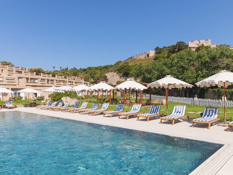 Klares Schwimmbecken mit Liegestühlen und Sonnenschirmen vor einem Hotel am Hang.