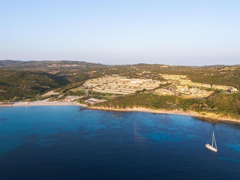 Küste mit blauem Meer und einem Segelboot, darüber Hügel mit Gebäuden unter klarem Himmel.