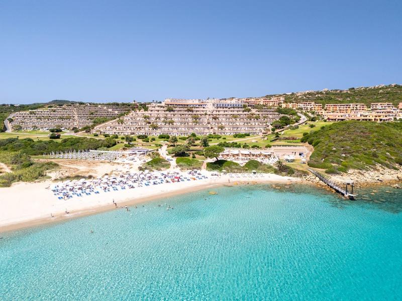 Panorama eines ruhigen Strandes mit klarem, türkisem Wasser und Hotelgebäuden im Hintergrund.