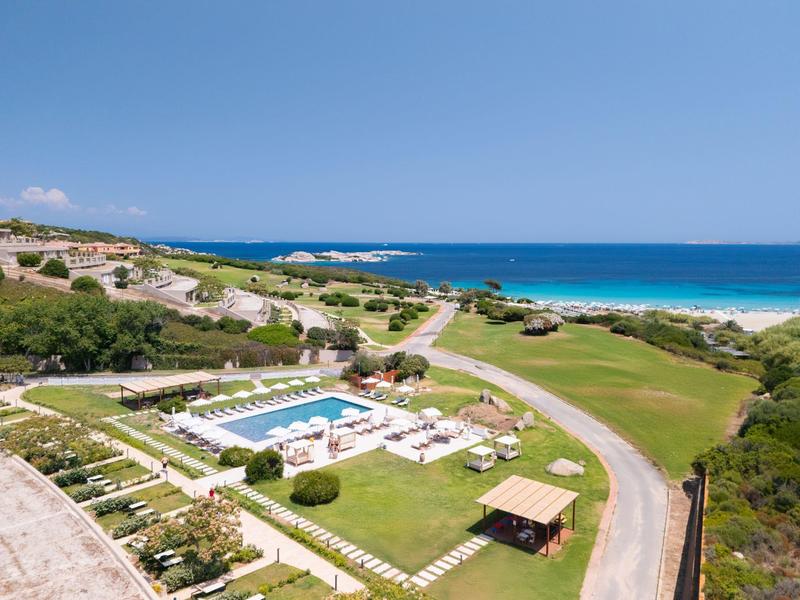 Blick auf einen luxuriösen Pool mit Liegestühlen, nahe einem Grasplatz und einem Sandstrand am Meer.