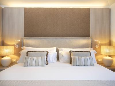 Moderní hotelový pokoj s velkou postelí, čelem postele, čtecími lampami a dvěma nočními stolky s lampami.