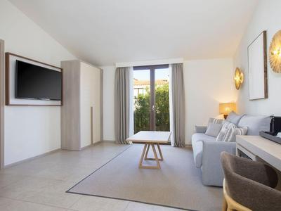 Světý obývací pokoj se šedou pohovkou, TV, jídelním stolem a velkými okny s výhledem do zeleně.