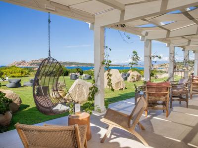 Zastřešená terasa s posezením a výhledem na zahradu a moře za jasné oblohy.