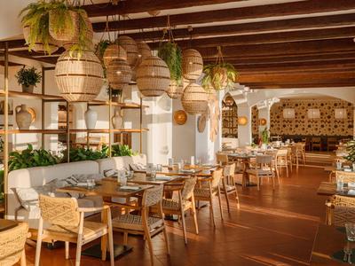 Světlá restaurace s dřevěným nábytkem, rostlinami a pletenými lampami pod viditelnými dřevěnými trámy.