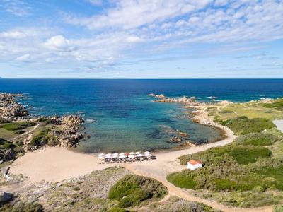 Playa de arena con sombrillas y agua clara, rodeada de arbustos verdes y rocas.