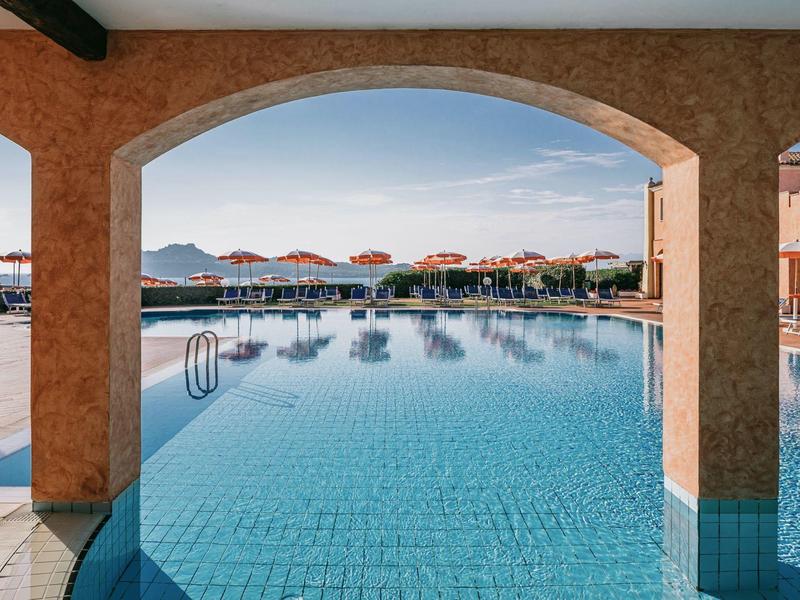 Pohled na venkovní bazén s modrými dlaždicemi, viděný z kryté terasy s oblouky.