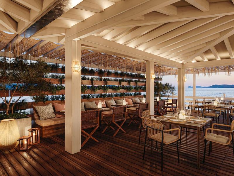 Stylová restaurace s dřevěnou podlahou, židlemi a svíčkami s výhledem na moře za západu slunce.