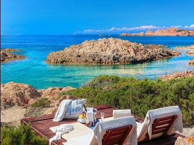 Zwei Sonnenliegen mit Handtüchern auf Holzdeck vor blauem Meer und Felsenlandschaft.