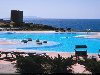 Buitenzwembad met ligstoelen uitkijkend over zee en een oude toren op de achtergrond.