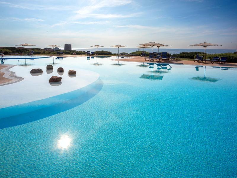 Groot buitenzwembad met ligbedden en parasols onder een zonnige hemel bij een resort.