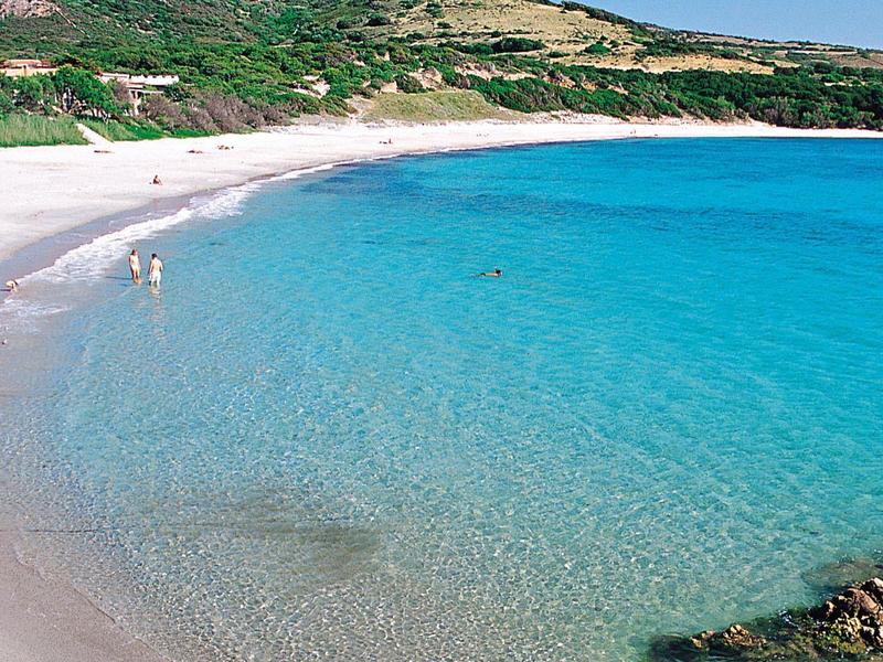 Helder turquoise water bij een gebogen wit zandstrand met groen in de achtergrond.
