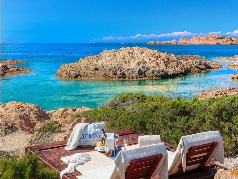 Twee ligstoelen bij rotsachtige kust met helderblauwe zee en eiland op de achtergrond.