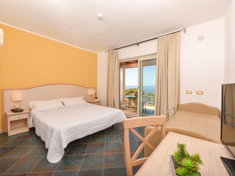 Camera d'hotel luminosa con letto matrimoniale, scrivania e vista su terrazza e mare.