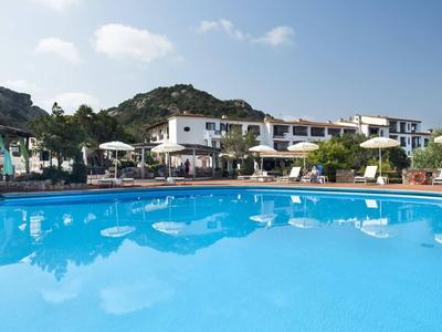 Großer Pool mit klarem blauem Wasser vor weißen Gebäuden und grünen Hügeln unter blauem Himmel.