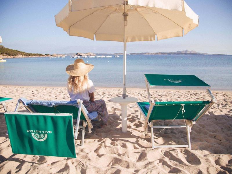 Donna in abito bianco con cappello da sole seduta sotto ombrellone sulla spiaggia accanto a lettini verdi.