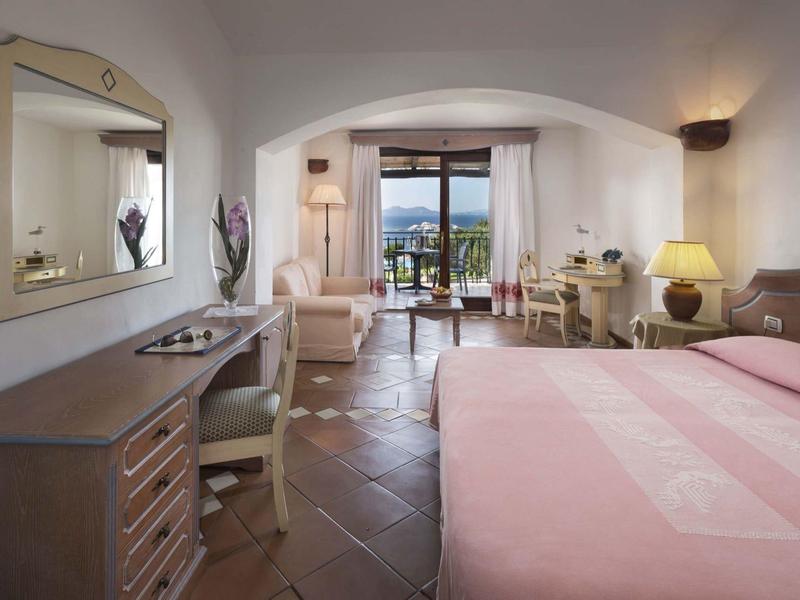 Camera d'hotel luminosa con letto matrimoniale, scrivania, sedie e vista su balcone e mare.