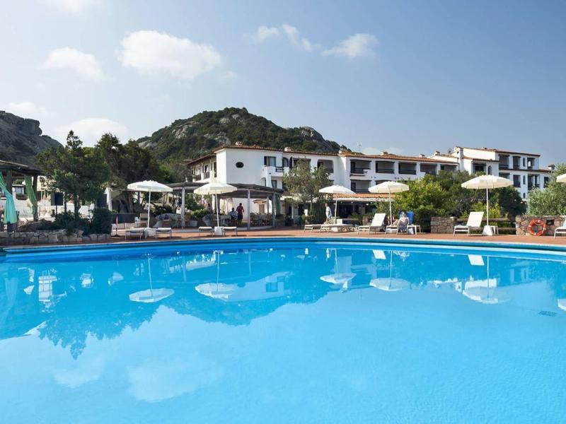 Grande piscina blu di fronte a un hotel con montagne sullo sfondo e cielo sereno.
