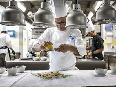 Kuchař v bílém oblečení zdobí pokrm v profesionální kuchyni pod velkými lampami.