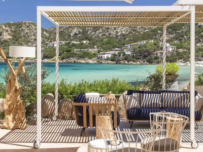 Outdoor-Sitzbereich mit Pergola, blauen Kissen, Holztisch und Blick auf türkisfarbenes Meer und Hügel.