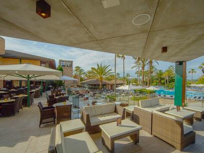 Lounge al aire libre con sillones y mesas junto a la piscina del hotel bajo sombrillas y palmeras.