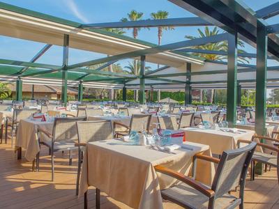 Área de restaurante al aire libre con mesas preparadas y vista a palmeras y piscina bajo sol.