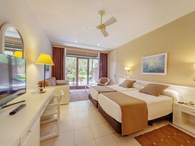 Habitación de hotel luminosa con dos camas, televisor, escritorio y acceso a un balcón con vista al jardín.