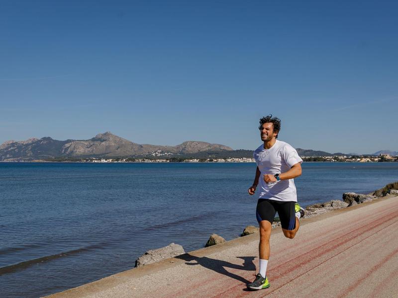 Hombre corre a lo largo de la costa por un camino soleado con mar y montañas al fondo.