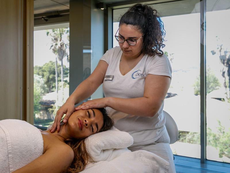 Una mujer recibe un masaje en cabeza y cuello en una sala de spa luminosa con vista al jardín.