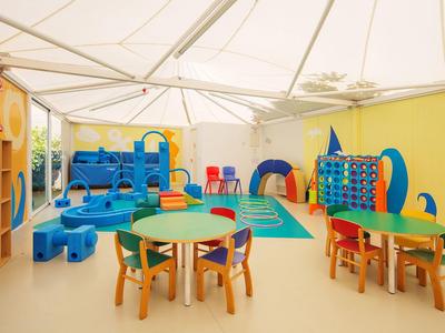 Heller, bunter Kindergartenraum mit Tischen, Stühlen, Spielhaus und Regalen unter weißem Zeltdach.