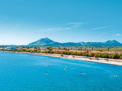 Klares blaues Meer vor Sandstrand mit Menschen und grüner Bergkulisse unter blauem Himmel.