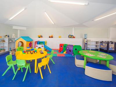 Heller Spielraum mit bunten Kindertischen, Stühlen, Spielhäusern und weichem blauen Boden.