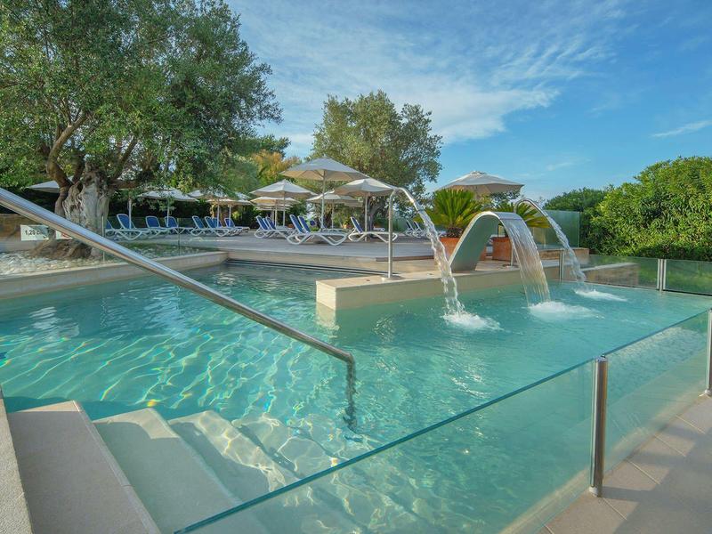 Piscine extérieure moderne avec fontaines, transats et parasols par une journée ensoleillée.