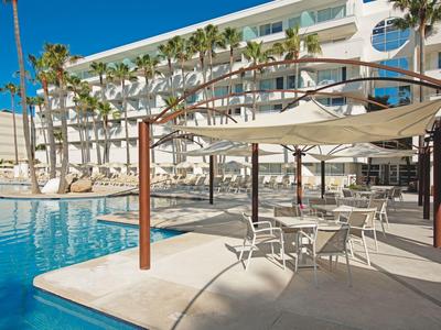 Area piscina moderna con ombrelloni, sedie e palme accanto a un edificio alberghiero bianco.