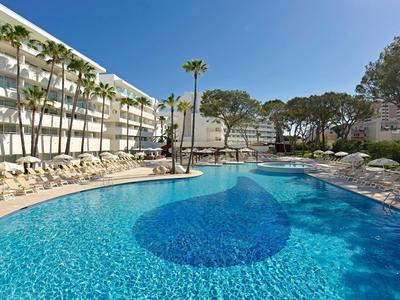 Grande piscina dell'hotel con acqua azzurra limpida, circondata da palme e lettini sotto un cielo sereno.