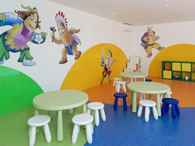 Salle de jeux colorée pour enfants avec petites tables, tabourets et fresques murales de personnages de dessin animé.