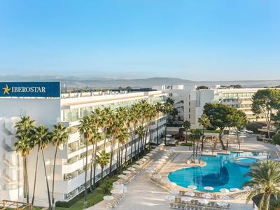 Hôtel avec piscine extérieure, entouré de palmiers et de bâtiments sous un ciel clair.