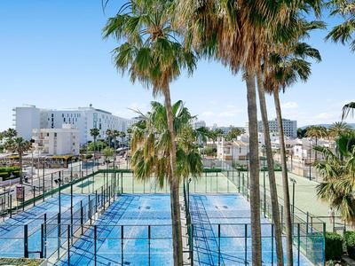 Vista de una pista de tenis azul con palmeras y edificios blancos al fondo bajo un cielo despejado.