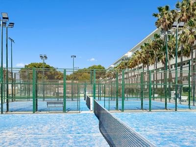 Terrains de tennis bleus à côté d'un bâtiment moderne avec des palmiers sous un ciel clair.