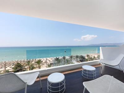 Balkon mit weißen Stühlen und Tischen, Glasgeländer, Blick auf Strand und blaues Meer.