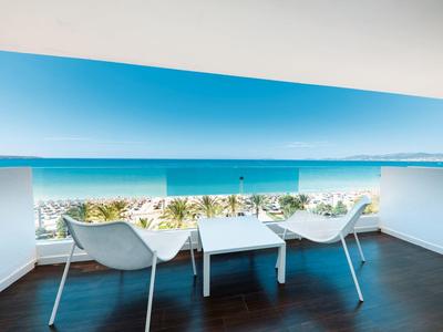 Balkon mit zwei weißen Stühlen, kleinem Tisch und Blick auf Meer sowie Palmen bei klarem Himmel.